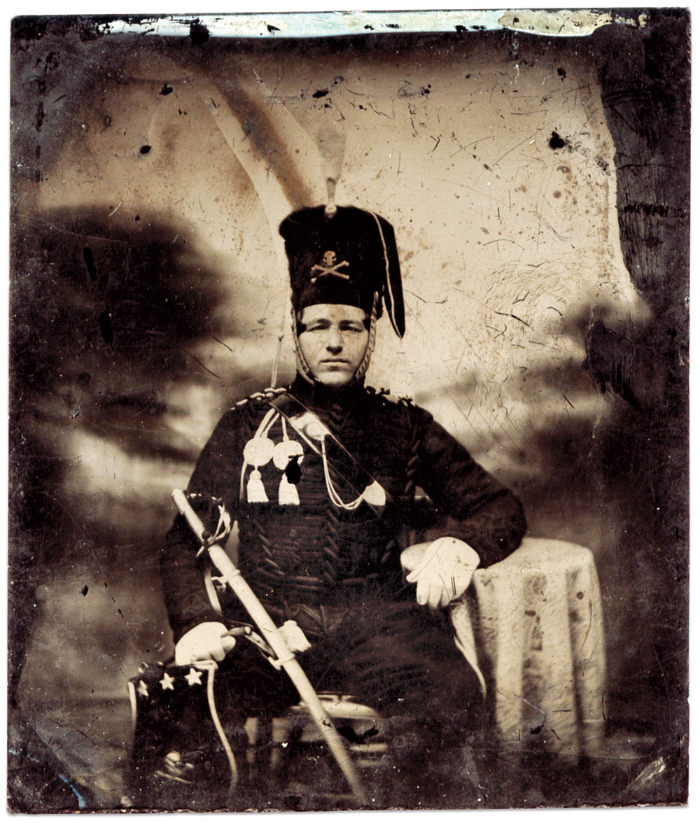 This American hussar, circa 1860, right, wears a uniform influenced by the European Black Hussars of the Napoleonic Wars. His fur-bodied hussar busby with brass-scaled chinstrap has a Totenkopf or death’s head skull and cross bones insignia attached, plus a white worsted pompon and hanging red cloth bag. His black jacket has a hussar-style braid with toggled ends across the chest. Although the baldric or patent leather sling with brass fittings, and aiguillette, or cord with lace tags, on his shoulder were usually worn by an adjutant or staff officer, most hussar officers appear to have worn them. A Model 1840 Dragoon saber made by the Ames Manufacturing Company rests on his leg, and a sabertache, or flat satchel, with three stars and another death’s head insignia, hangs from long straps from his sword belt. This image appeared on the cover of the July-August 1986 issue of MI. Sixth plate magenta ambrotype by an unidentified photographer. Author’s collection. 