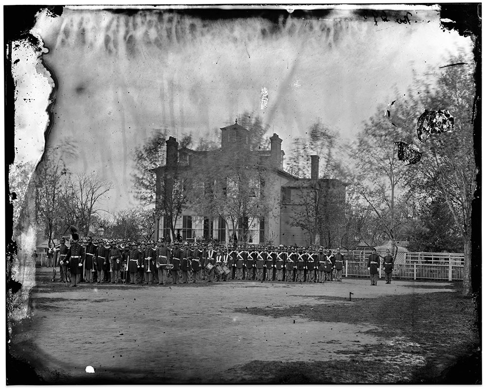 This circa 1864 view of the Marine Band in front of a company from the Marine Barracks in Washington, D.C., with the Commandant’s house in the background has been frequently published. The company commander stands to the right and is next to the sergeant major, who is identifiable by his uniform’s gold lace. Sergeants in the formation carried the cartridge box, bayonet, and sword from a waist belt while corporals and privates continued using shoulder belts. The enlisted uniform of Navy blue, double-breasted, edged red, had yellow loops to the collar and a slashed cuff. Musicians had scarlet coats, trimmed with yellow lace to the collar and cuffs, though technical limitations of this period show the bright yellow as a much darker tone, so the lace is difficult to see, but their placement is identifiable by their bright buttons. Glass plate negative by an unidentified photographer. Library of Congress.