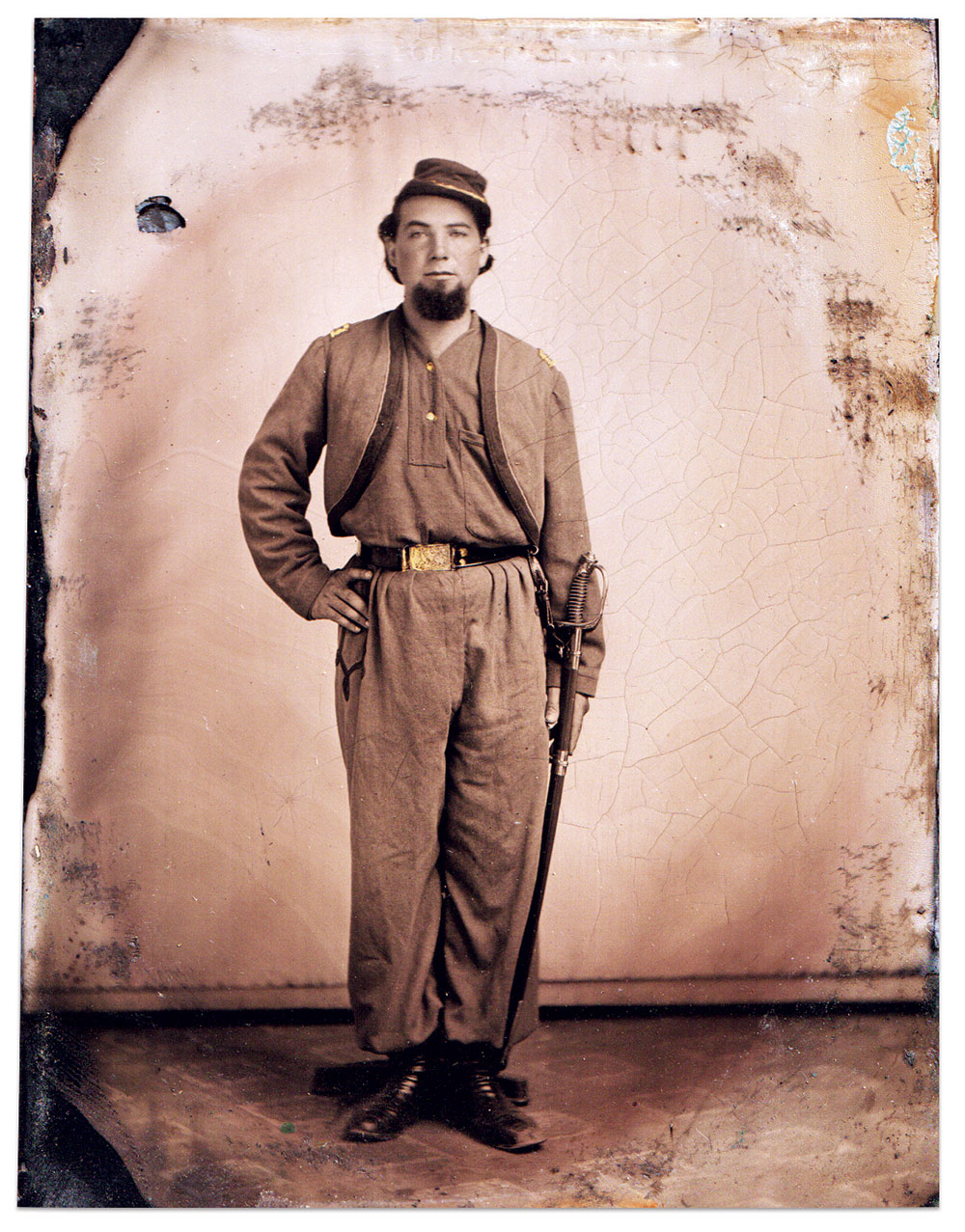 The uniform acquired by this unidentified officer of Wallace’s Zouaves in May 1861 featured the red trim edged with light blue. A small modified “Austrian” knot is visible on his chasseur-pattern trousers. His officer’s kepi is red overall. His collarless blue flannel shirt has a placket front with two buttons, and a large patch breast pocket. His waist belt is being worn upside down in order that his sword may appear as if it is being worn on the correct side in his reversed image. Half-plate tintype by an unidentified photographer. Dan Miller Collection.